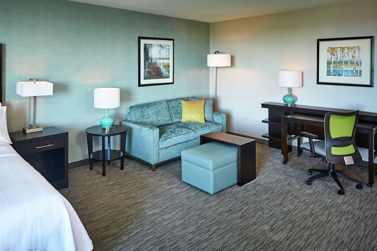 Ein Hotelzimmer mit einem Bett, einem Sofa, einem Schreibtisch und einem Sessel. Auf dem Schreibtisch stehen zwei Lampen und neben dem Sofa ein kleiner Tisch.