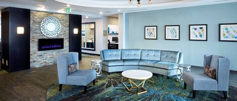 A modern lounge area with a curved grey sofa, two armchairs, a round coffee table, and a patterned rug. A fireplace with a TV screen is mounted on a brick wall, and there are framed artworks on the wall.