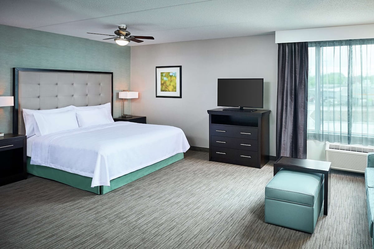 Ein Hotelzimmer mit Bett, Fernseher, Sofa, Schreibtisch und Deckenventilator.