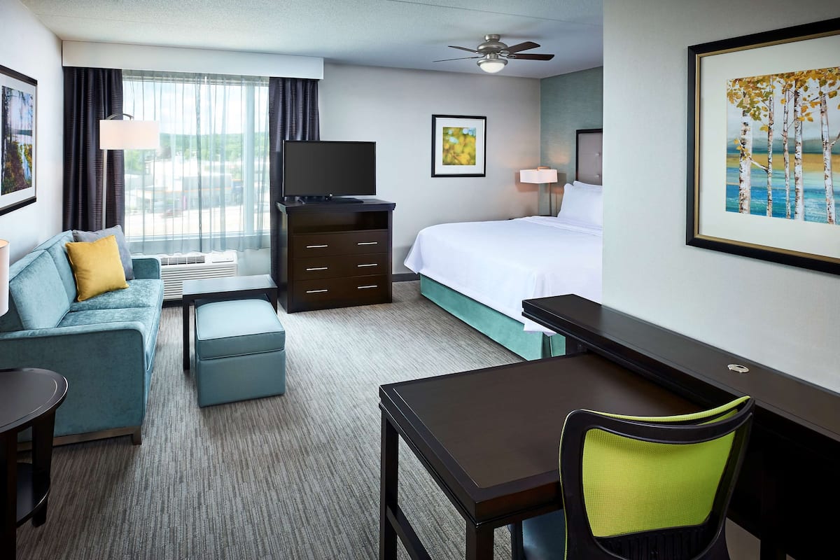 Ein Hotelzimmer mit Bett, einer Sitzecke, Schreibtisch, Stuhl, Fernseher und einem fenster mit Vorhängen.