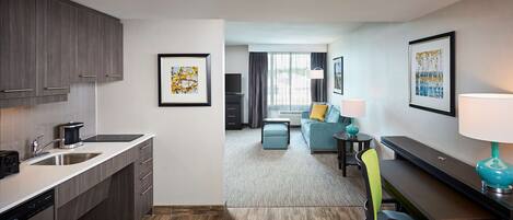 Una habitación de hotel moderna con kitchenette, sala de estar con sofá y televisor, y comedor con mesa y sillas.