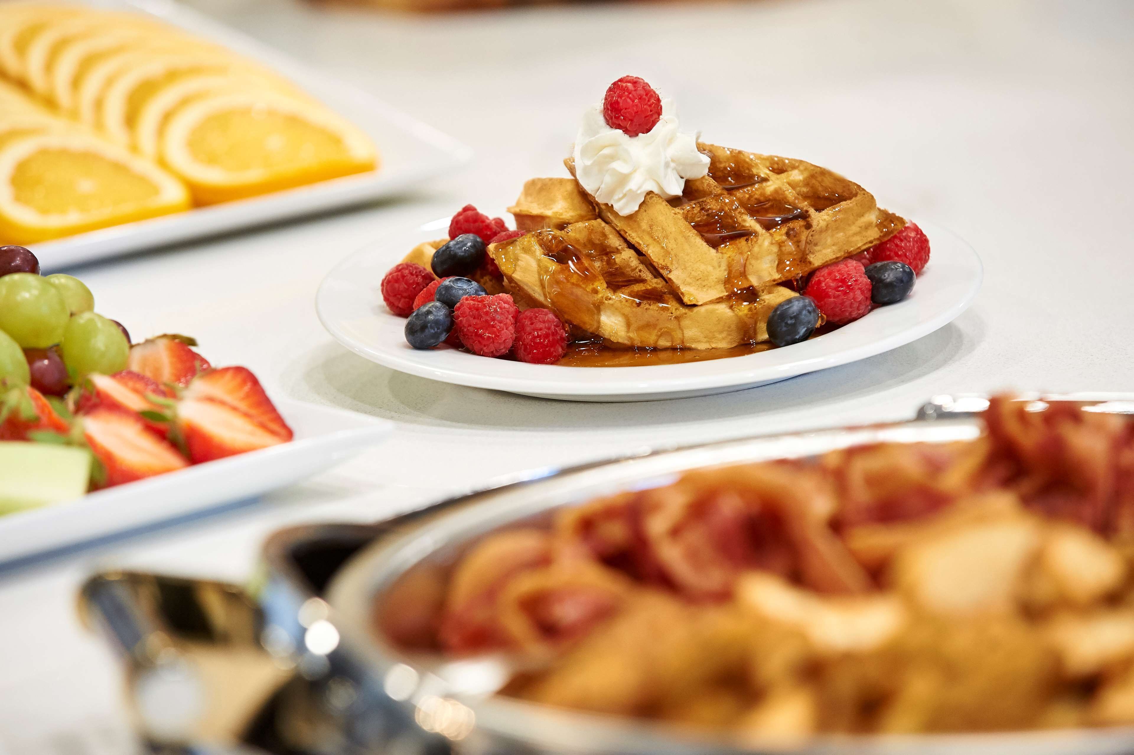 A breakfast buffet with waffles topped with whipped cream and berries, alongside sliced oranges and a variety of fruits.