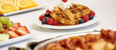 Un buffet de petit-déjeuner proposant des gaufres garnies de crème fouettée et de fruits rouges, ainsi que des tranches d’orange et une variété de fruits.