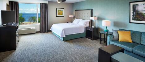 Habitación de hotel con cama, escritorio con televisor, sofá, mesita y ventana con vista al mar.