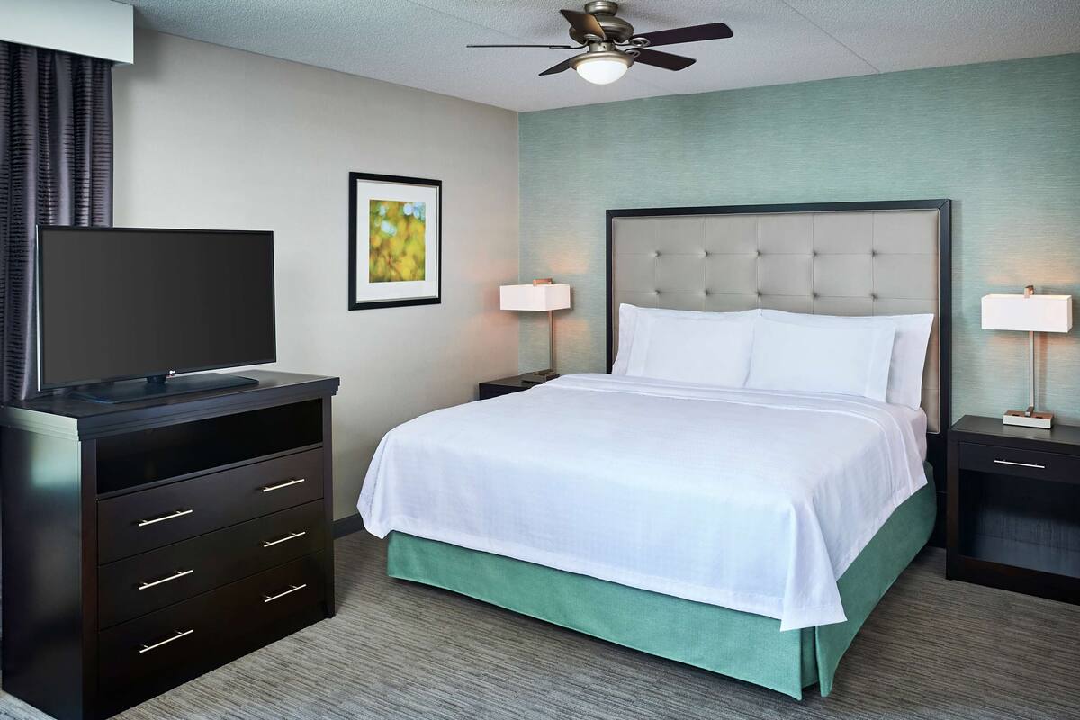 Ein Hotelzimmer mit einem Bett, einem Fernseher auf einer Kommode, zwei Nachttischen mit Lampen, einem Deckenventilator und einem Fenster mit Vorhängen.