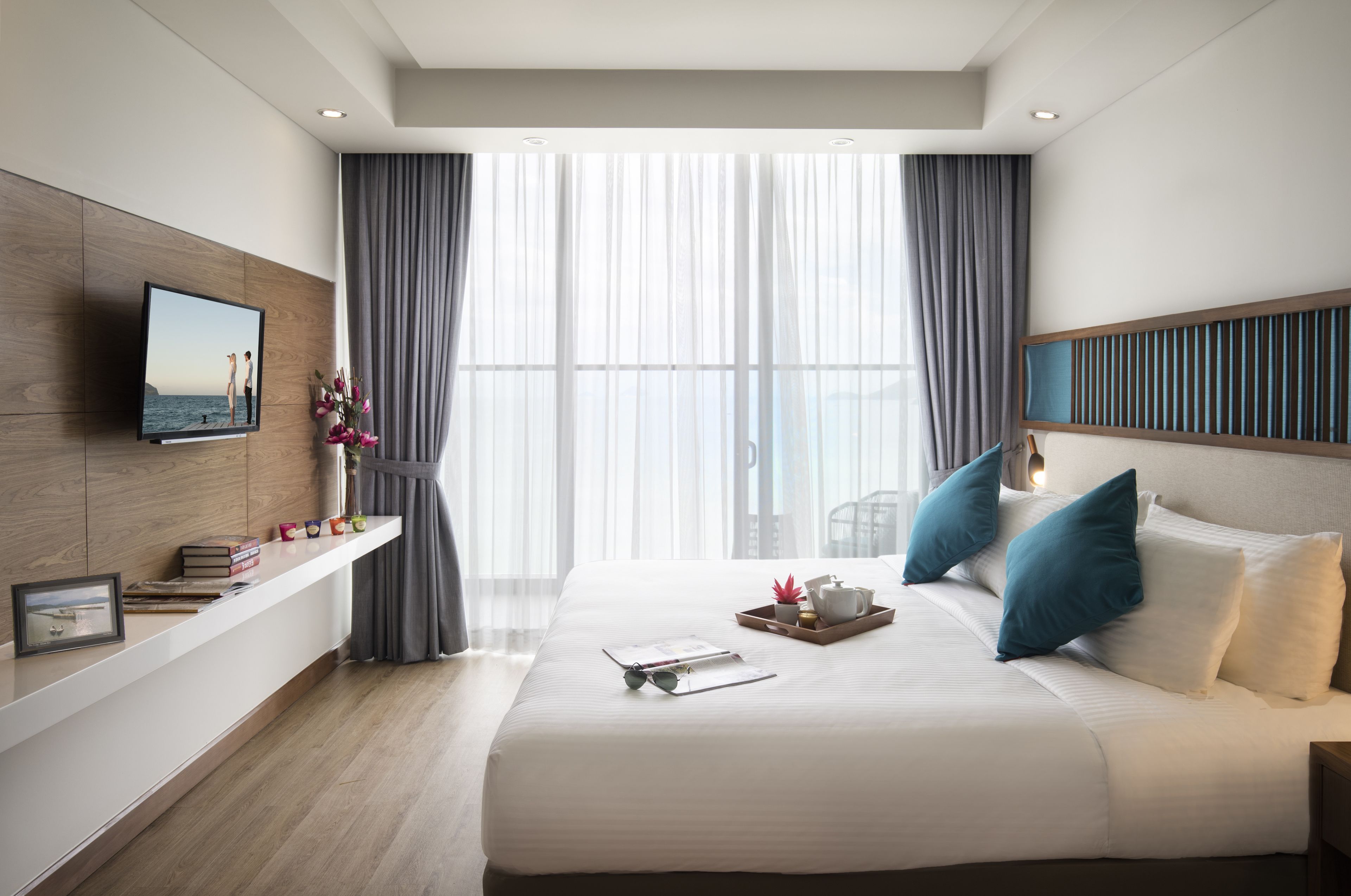 Una moderna camera d'hotel con un letto grande, una TV a parete, un comodino con libri e una cornice, e vista sul mare attraverso tende leggere.