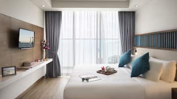Una moderna camera d'hotel con un letto grande, una TV a parete, un comodino con libri e una cornice, e vista sul mare attraverso tende leggere.
