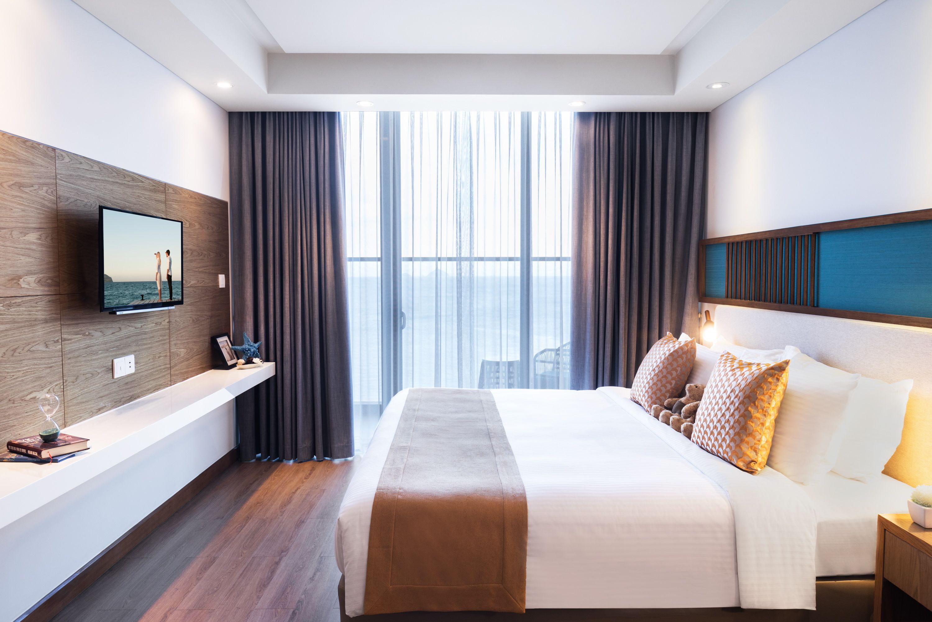 A modern hotel room with a large bed, a mounted TV, a bedside table with a book and a decorative item, and a view of the sea through large windows.