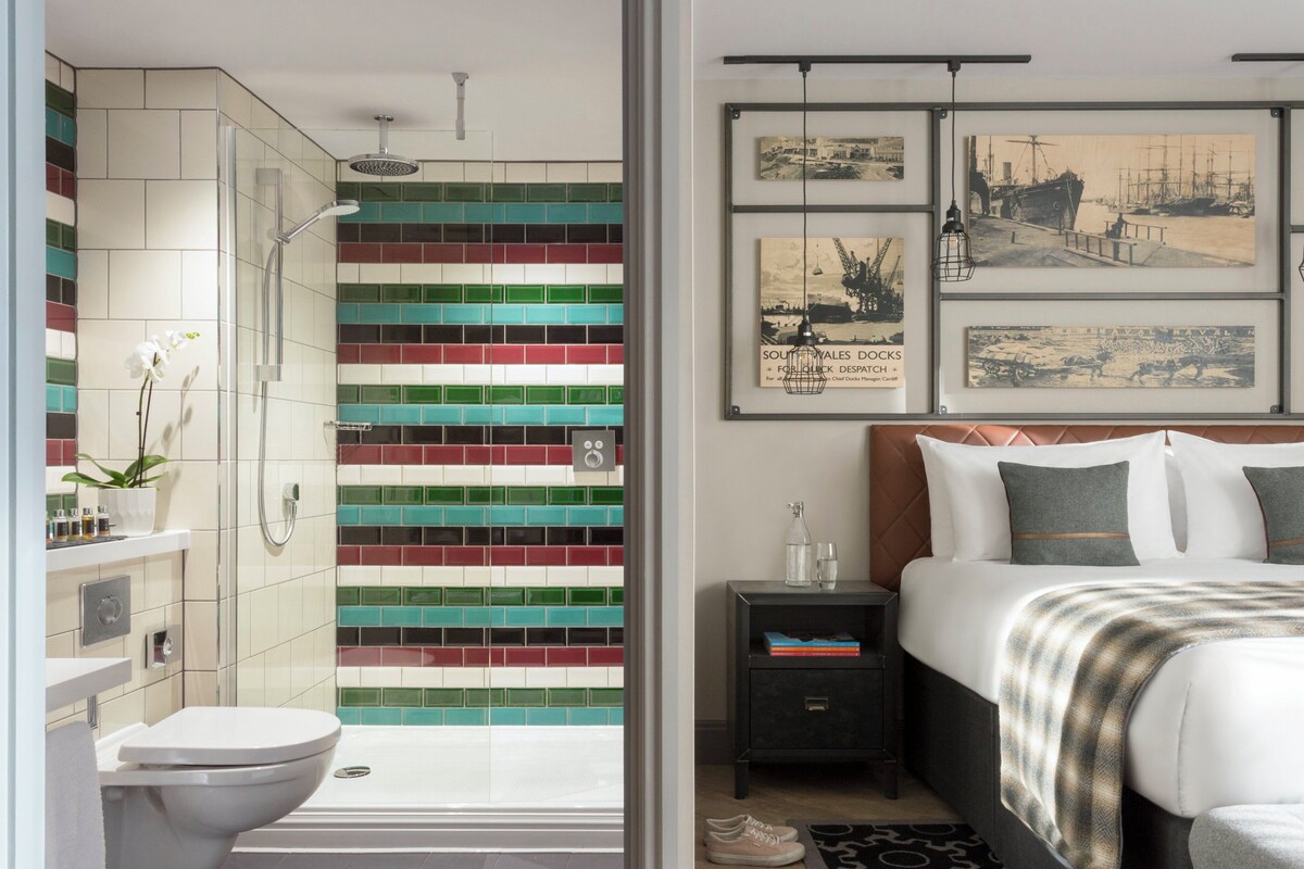 Un bagno moderno con doccia, water e parete rivestita di piastrelle. Si nota un letto con testiera e cuscini, e un comodino con lampada.