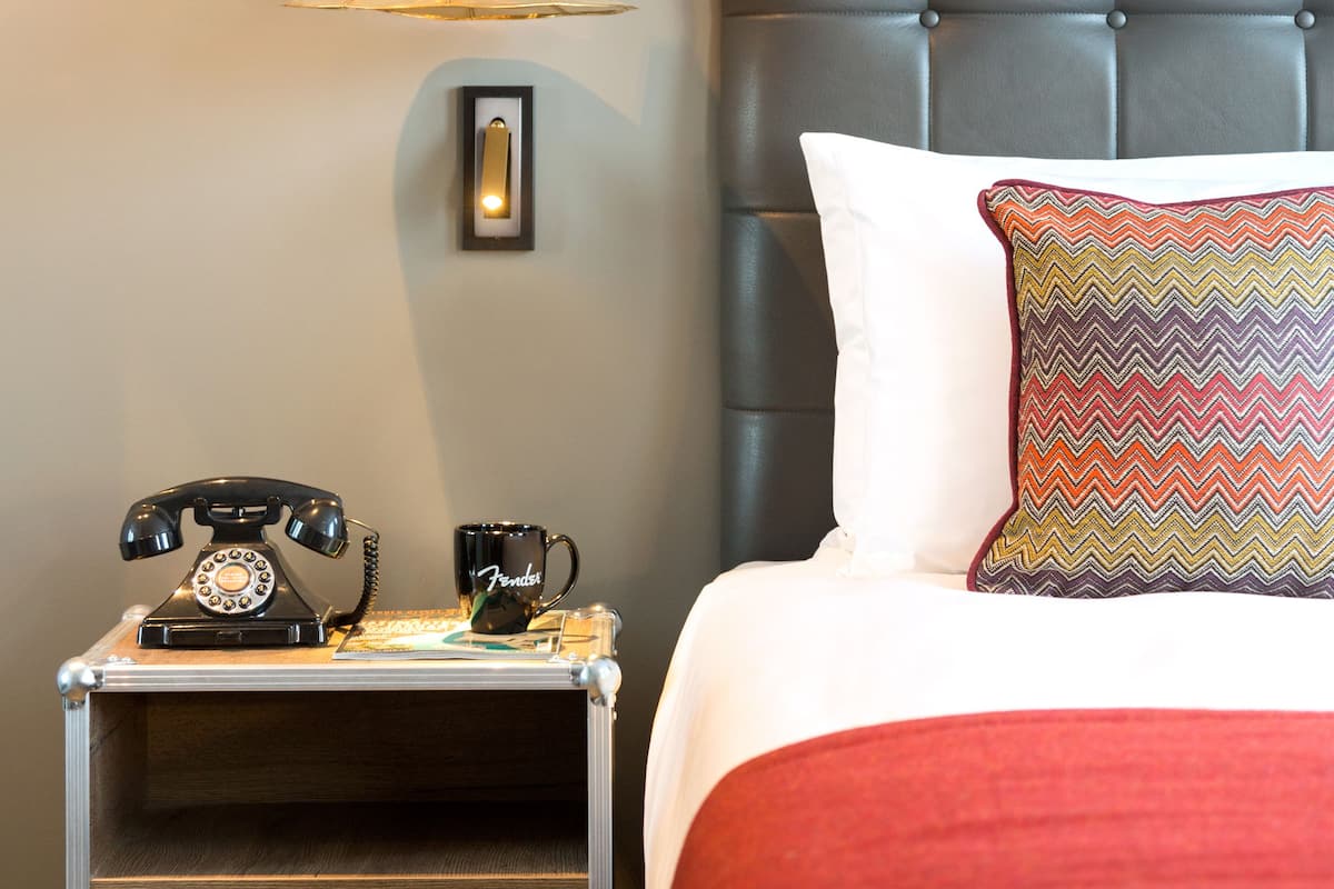 A neatly arranged hotel room with a bed, a bedside table with a telephone, a lamp, and a wall-mounted speaker system.
