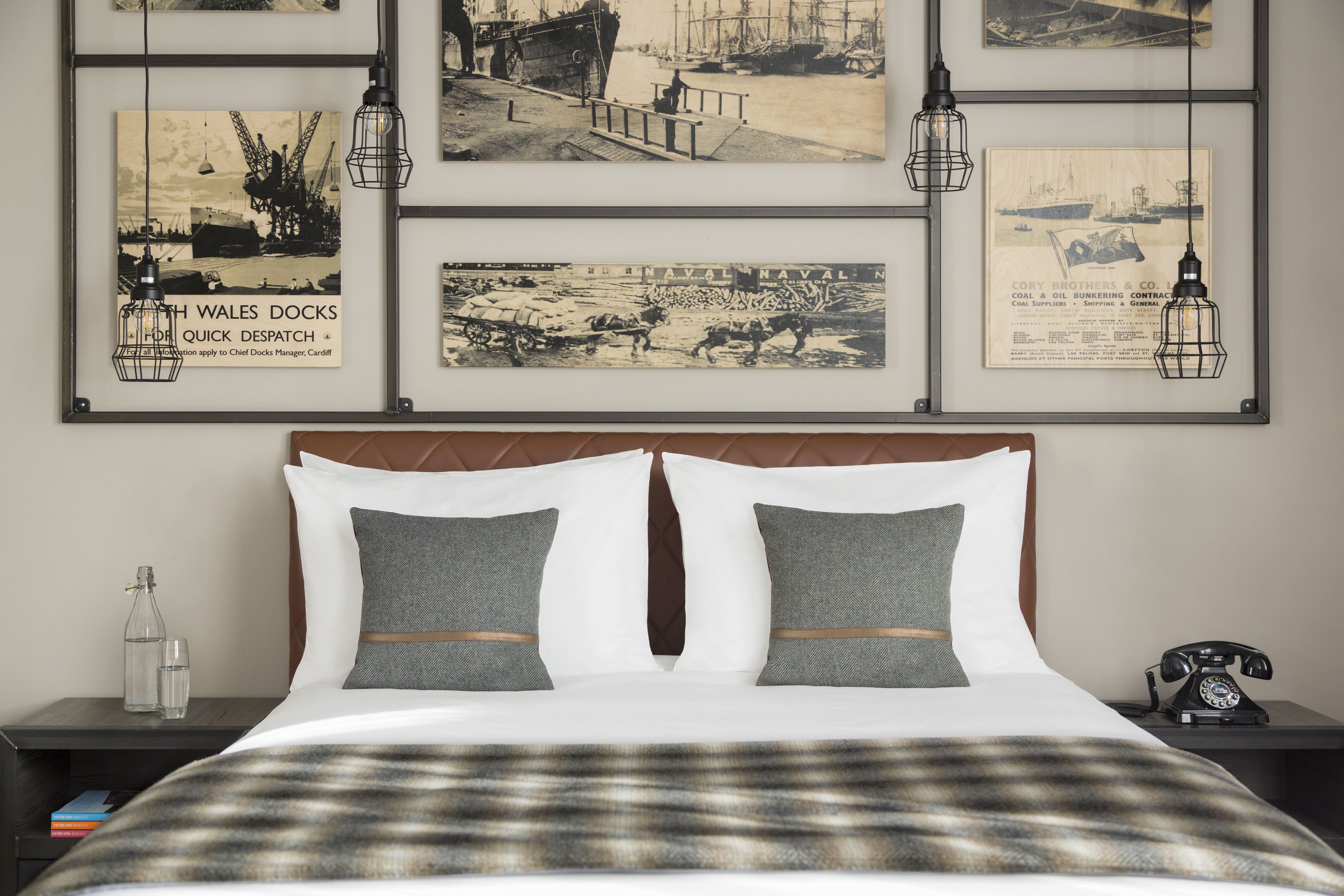 Een net opgemaakt bed met een geruite deken, twee grijze kussens en een houten hoofdbord. Boven het bed hangen foto's in lijsten en op het nachtkastje ernaast staat een telefoon.
