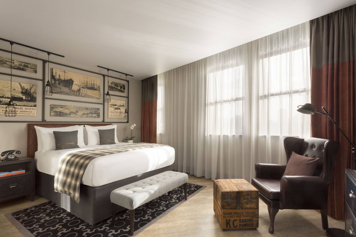 A modern hotel room with a large bed, a sitting area with a chair, a desk, and a box of K-C brand baking powder.