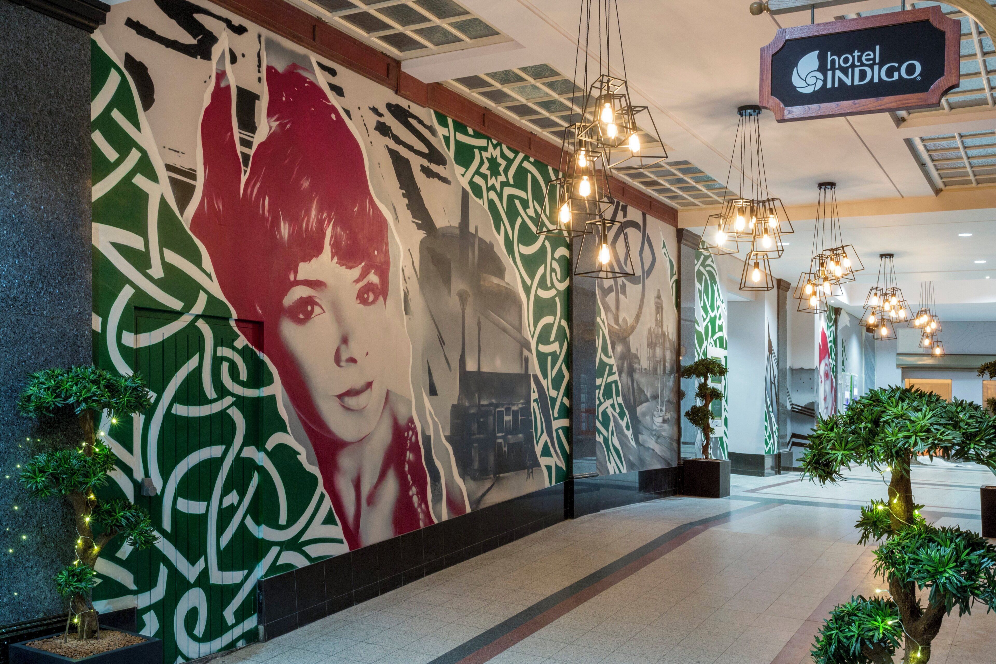 A hotel lobby with a large mural, modern lighting fixtures, and potted plants.
