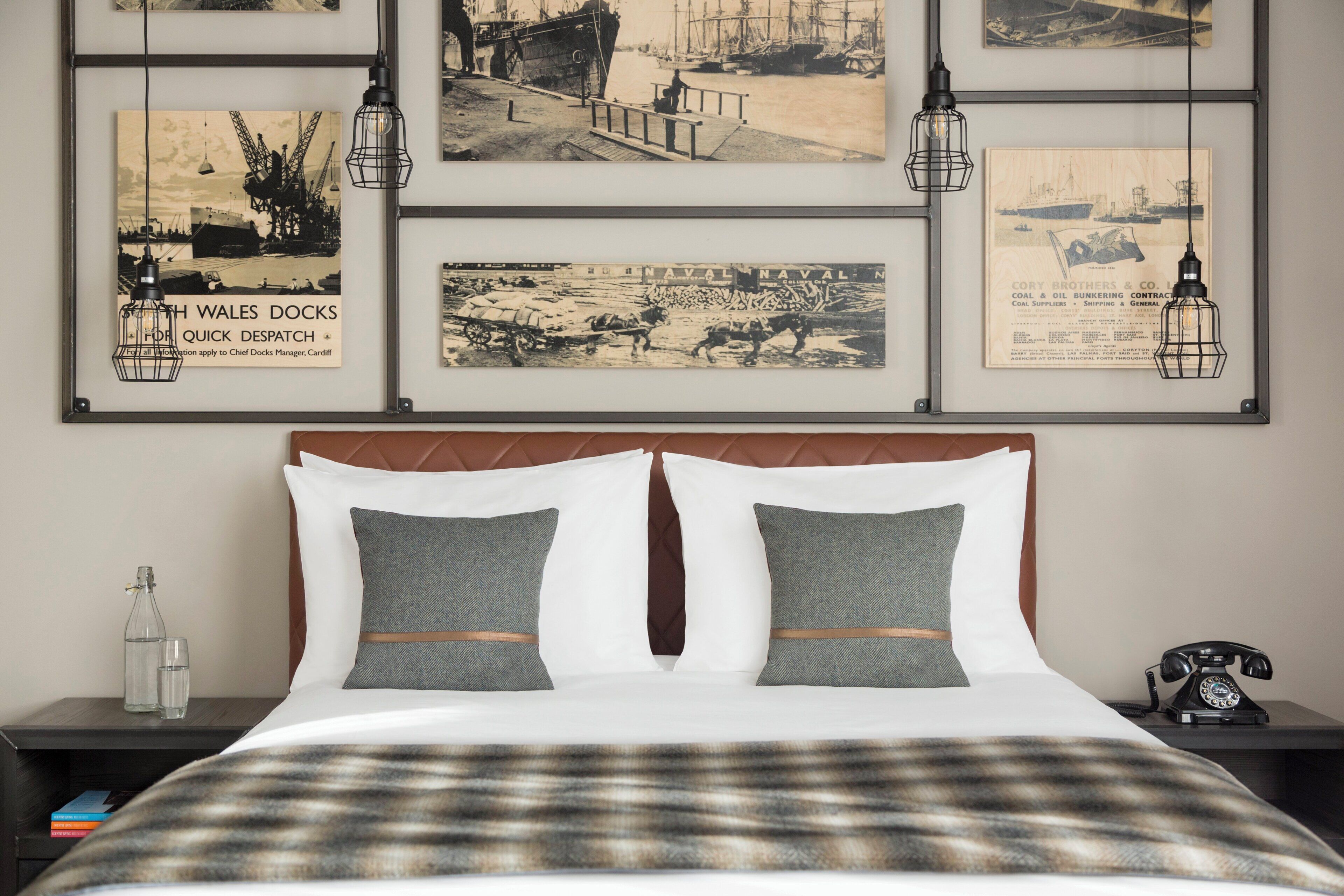 A hotel room with a bed, two pillows, a nightstand with a bottle and glasses, a telephone, and a wall with framed historical photographs.