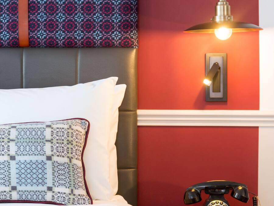 A neatly made bed with patterned pillows, a red wall, a vintage telephone, and a pendant light.