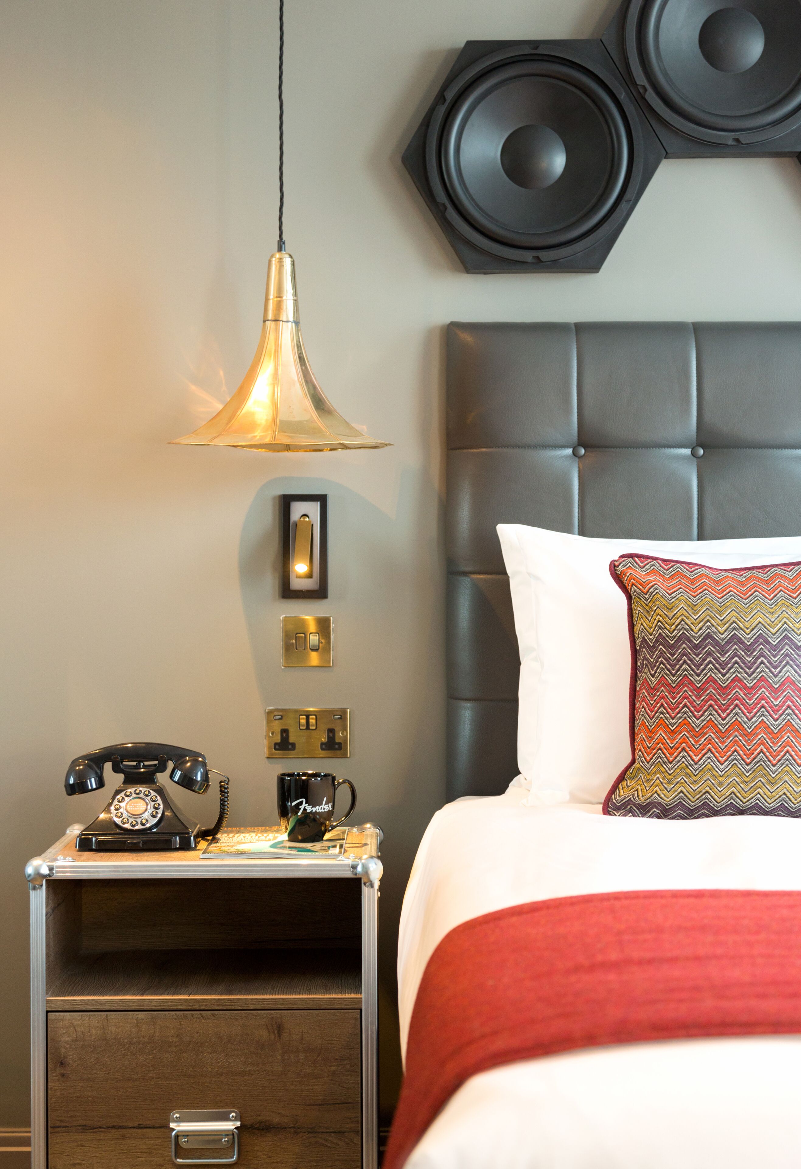 A neatly arranged hotel room with a bed, a bedside table with a telephone, a lamp, and wall-mounted light switches.