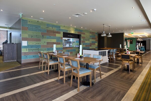 Um restaurante moderno com mesas e cadeiras de madeira, área de cozinha e parede verde com design padronizado.