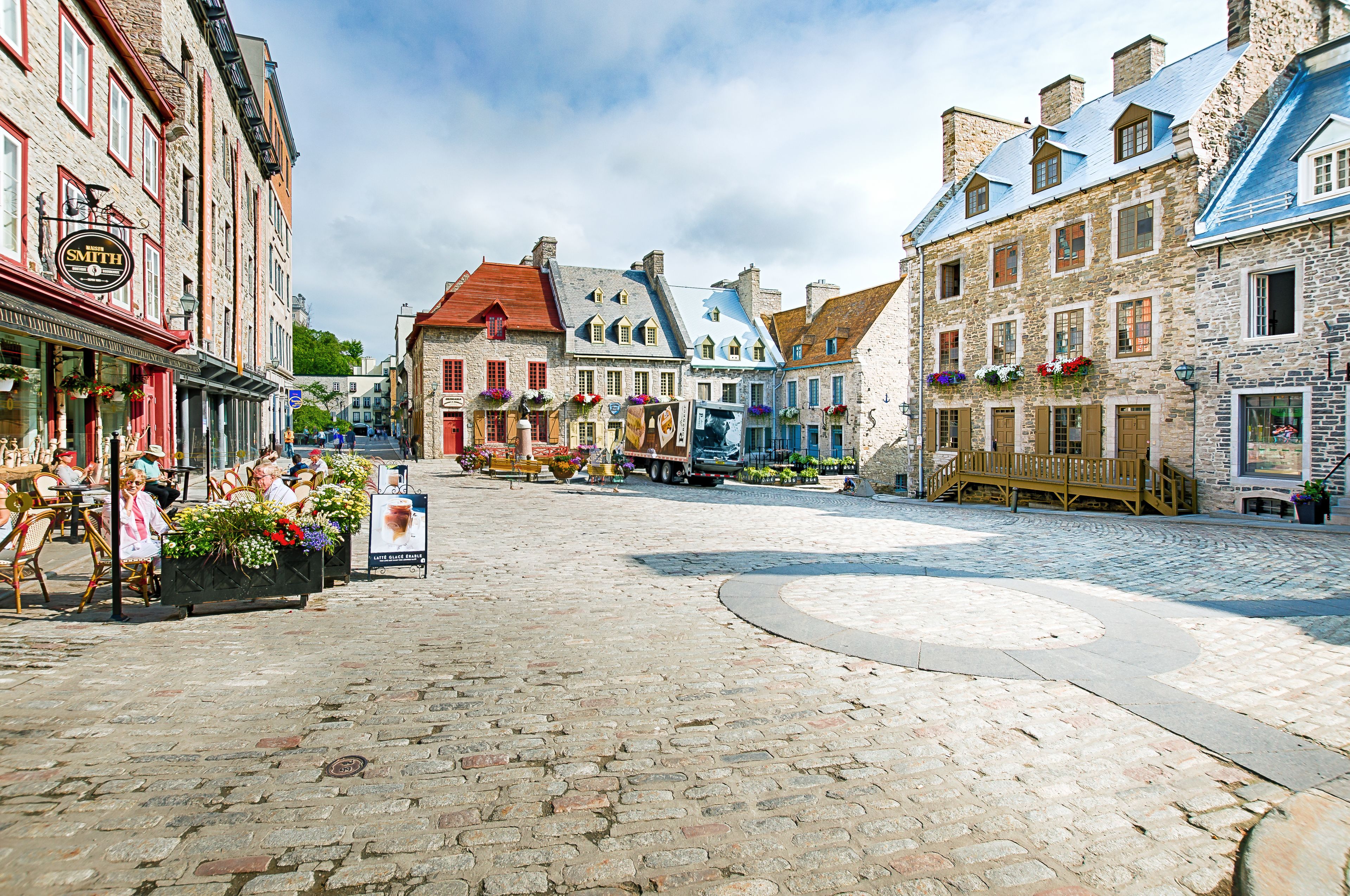 Ein Kopfsteinpflasterplatz mit historischen Gebäuden, Außenbestuhlung und Blumenarrangements.