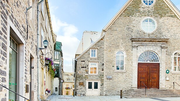 Una calle empedrada con edificios históricos de piedra, una iglesia con torre del reloj y un banco adornado con plantas en macetas.