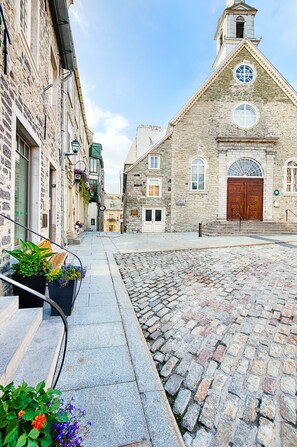 A cobblestone street with historic stone buildings, a church with a clock tower, and a bench with potted plants. - Les Lofts Champlain - By Les Lofts Vieux-Quebec (Québec City)