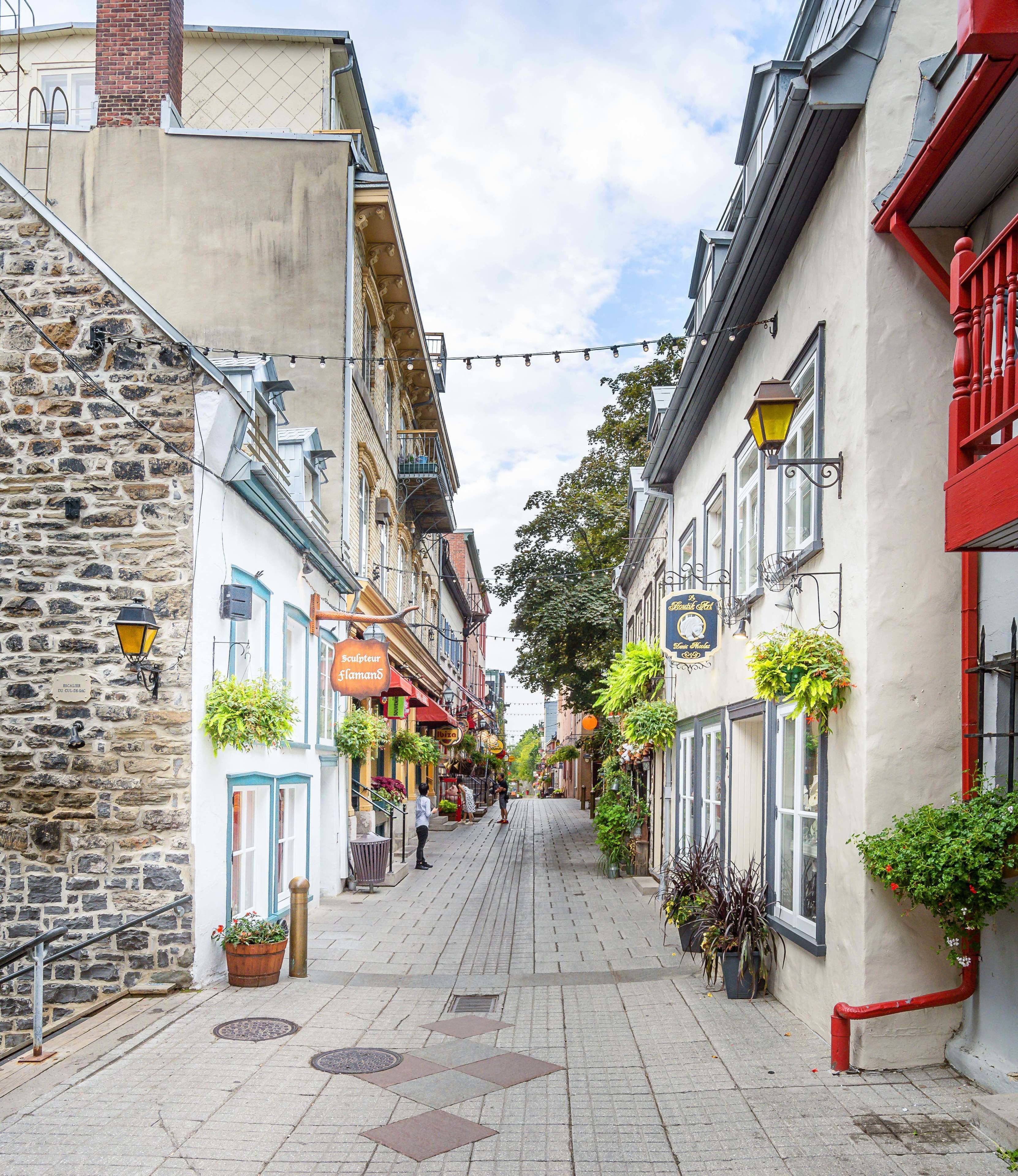 A narrow street lined with buildings, some with balconies and hanging plants.