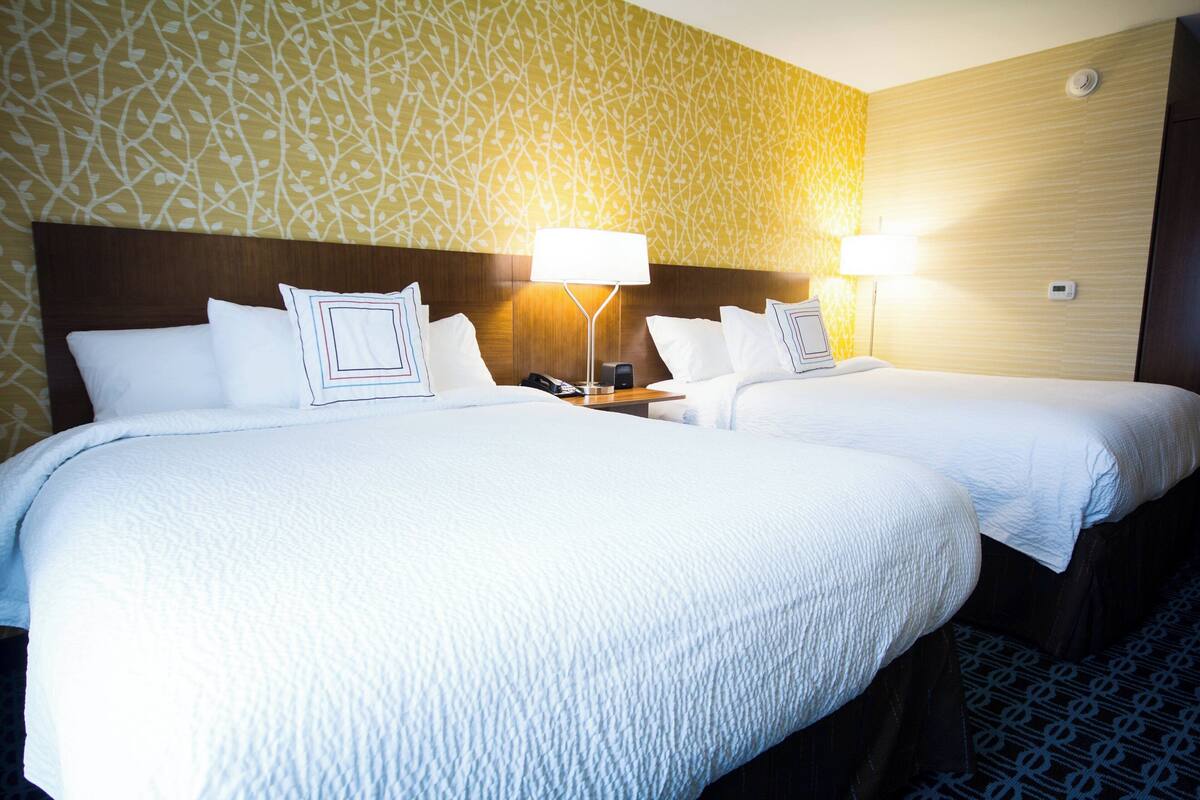 A hotel room with two beds, each with a white bedspread and matching pillows. There are two bedside tables with lamps, and the walls have a patterned wallpaper.