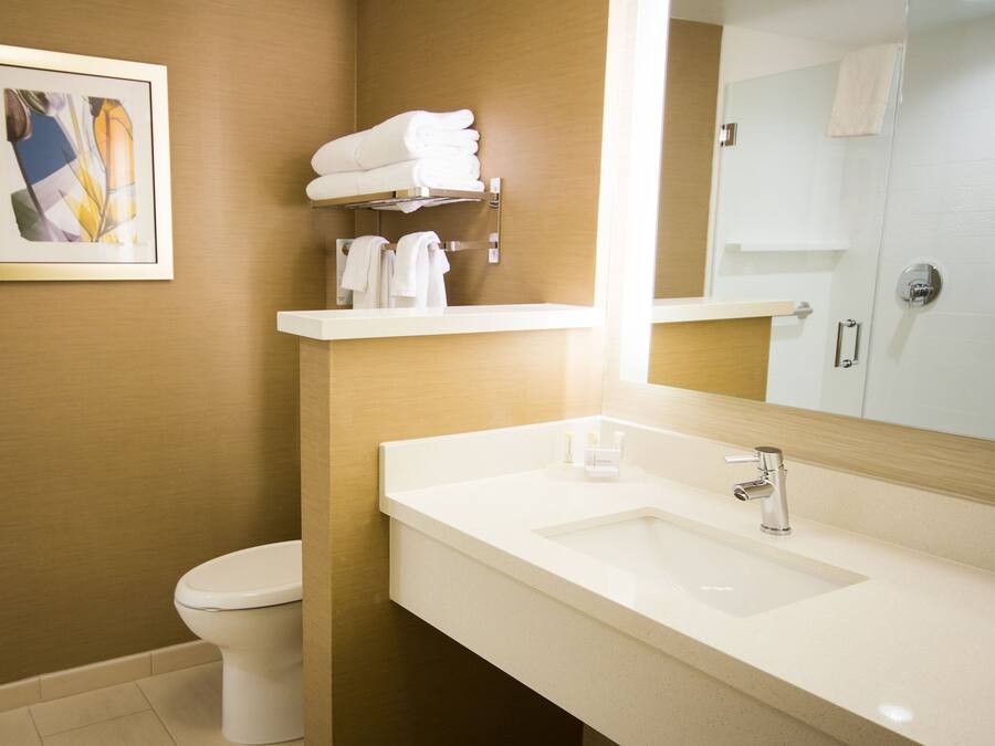 Ein modernes Badezimmer mit einem weißen Waschbecken, einem Spiegel und einer Toilette. Handtücher hängen auf einem Handtuchhalter, und an der Wand hängt ein gerahmtes Bild.