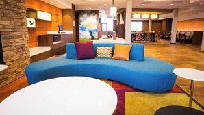 A modern hotel lobby with a curved blue sofa, a reception desk, and a seating area with tables. - Fairfield Inn and Suites Denver Northeast Brighton (Brighton)