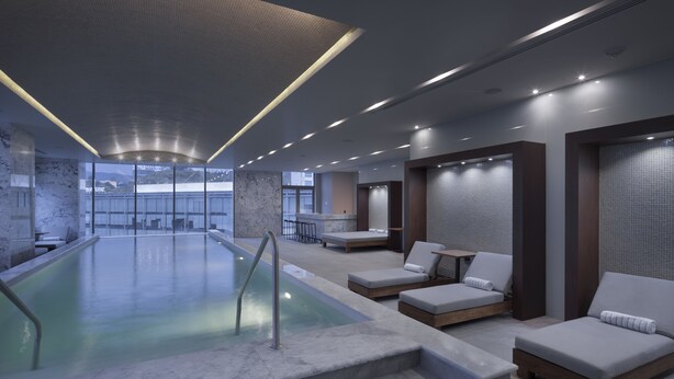 Un área de piscina interior moderna con vistas despejadas al exterior, un espacio de descanso con sillones reclinables y un ambiente tipo spa.