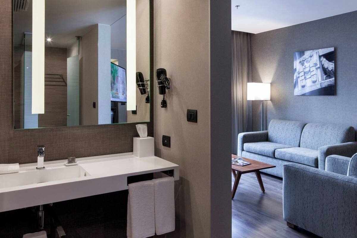 Une salle de bain d’hôtel moderne, dotée d’un grand miroir, d’un lavabo blanc et d’un revêtement de sol en bois foncé.