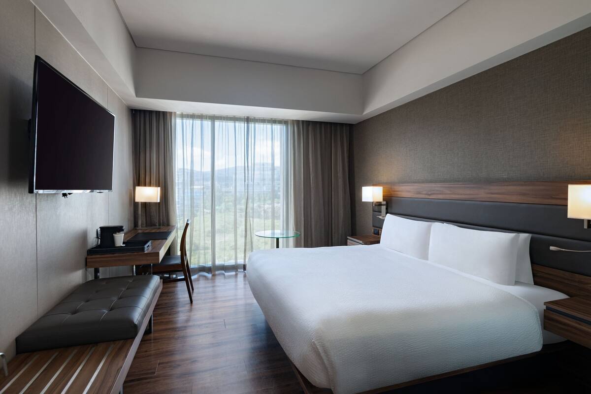 Une chambre d’hôtel dotée d’un grand lit, d’un téléviseur à écran plat, d’un bureau avec une chaise, d’une petite table et offrant une vue sur la ville par la fenêtre.