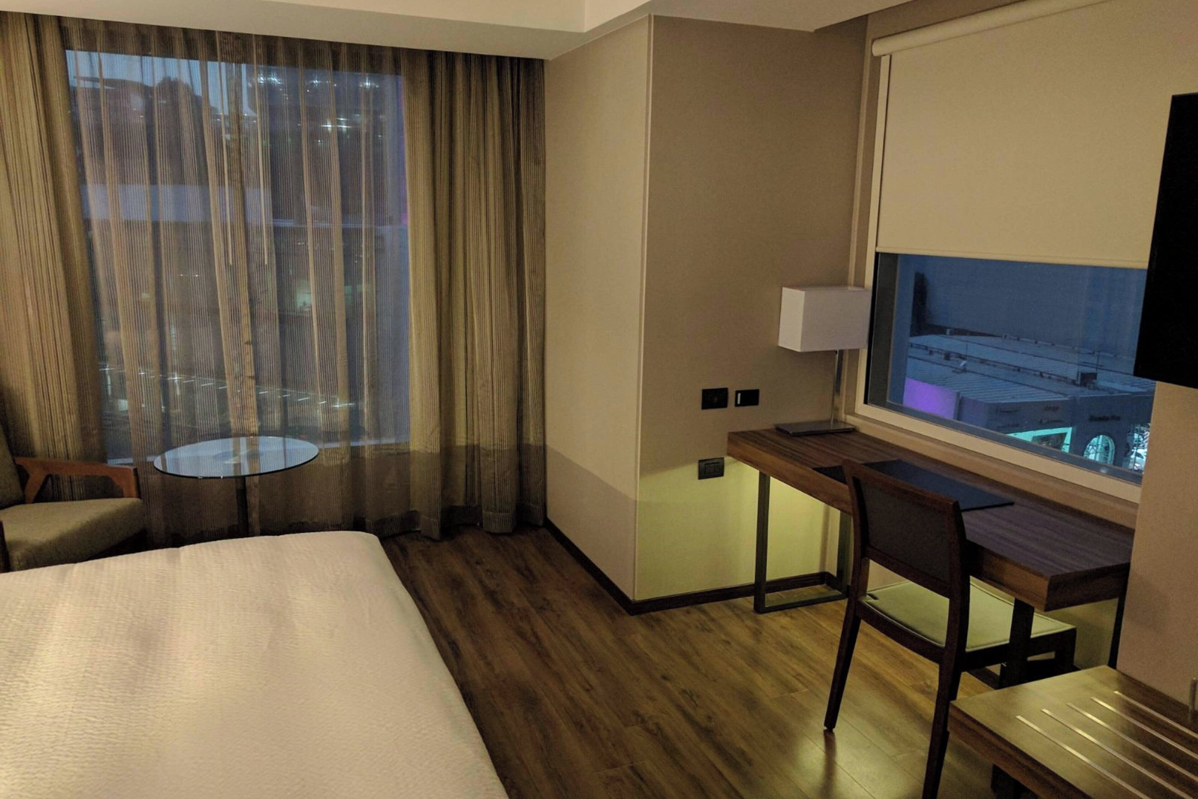 A hotel room with a bed, a desk with a chair, a small round table, and a window with curtains.