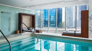 Piscina interior moderna con paredes de vidrio transparente, vistas a altos edificios y una zona de estar con sillones reclinables.