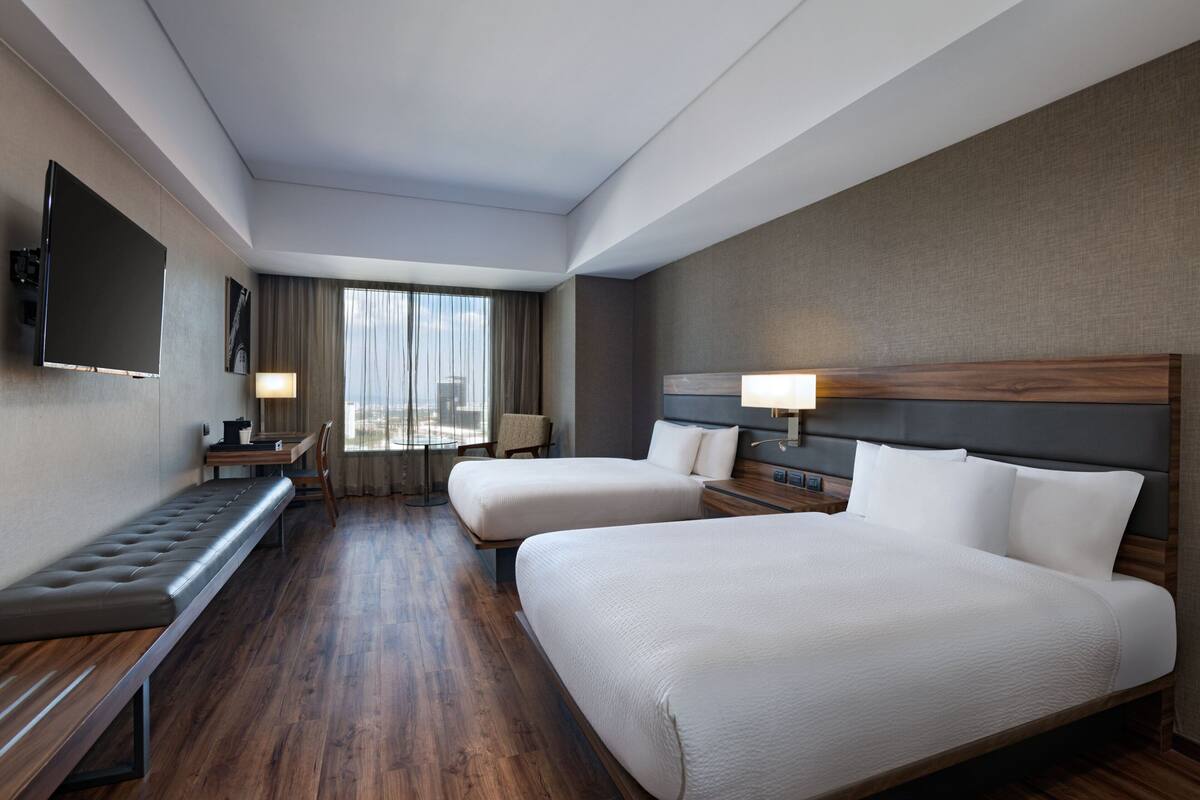 A hotel room with two beds, a bench, a desk, and a TV.