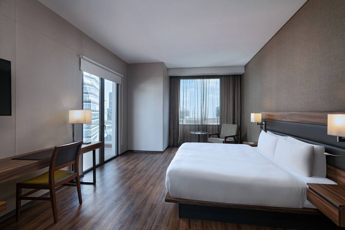 A modern hotel room with a large bed, a desk with a chair, a small table, and a view of the city through large windows.