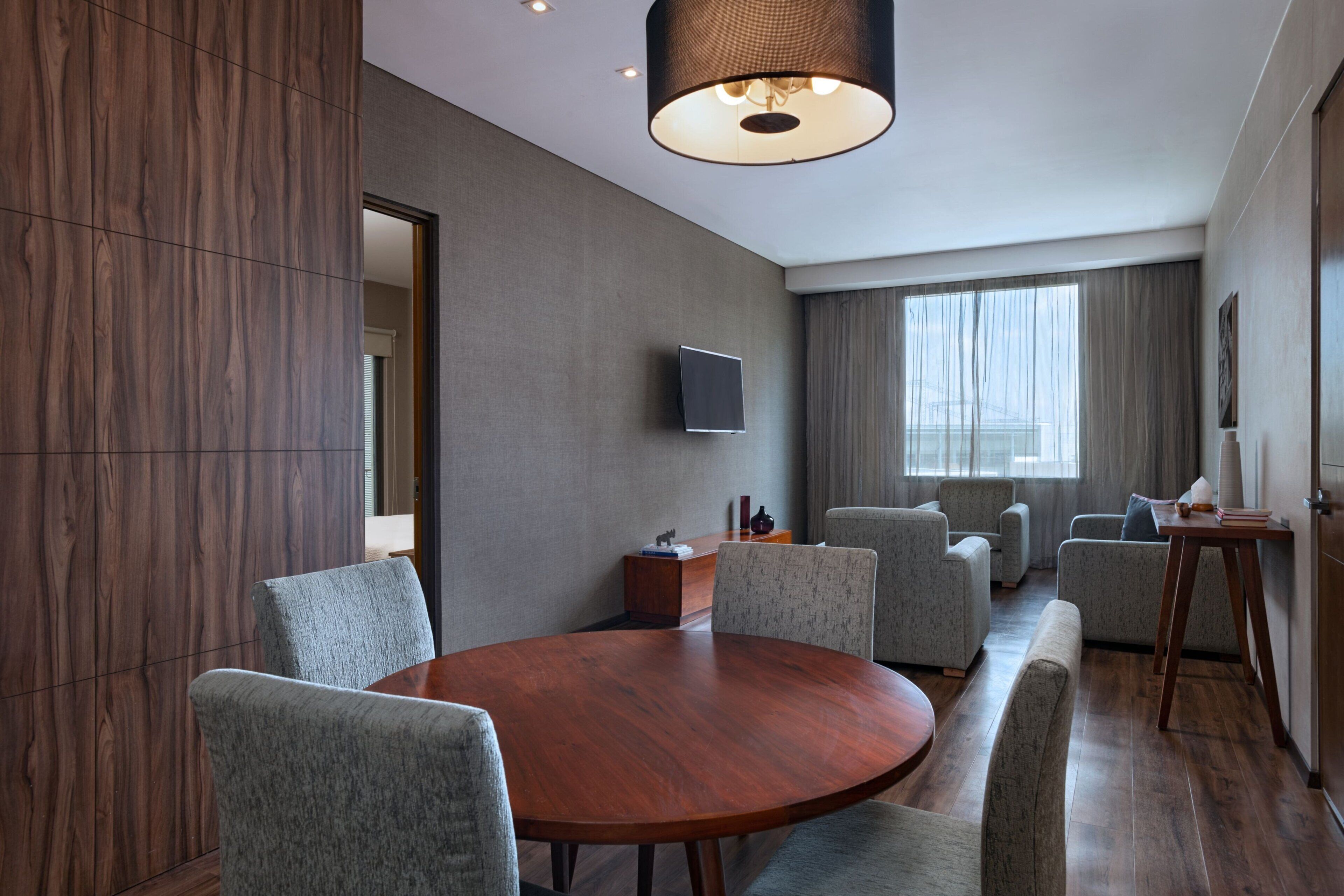 A modern conference room with a round wooden table, grey upholstered chairs, a flat-screen TV mounted on the wall, and a large window with curtains.