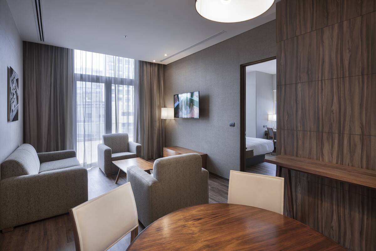 Une chambre d’hôtel moderne avec une table à manger en bois, deux canapés, un téléviseur et une grande fenêtre avec des rideaux.
