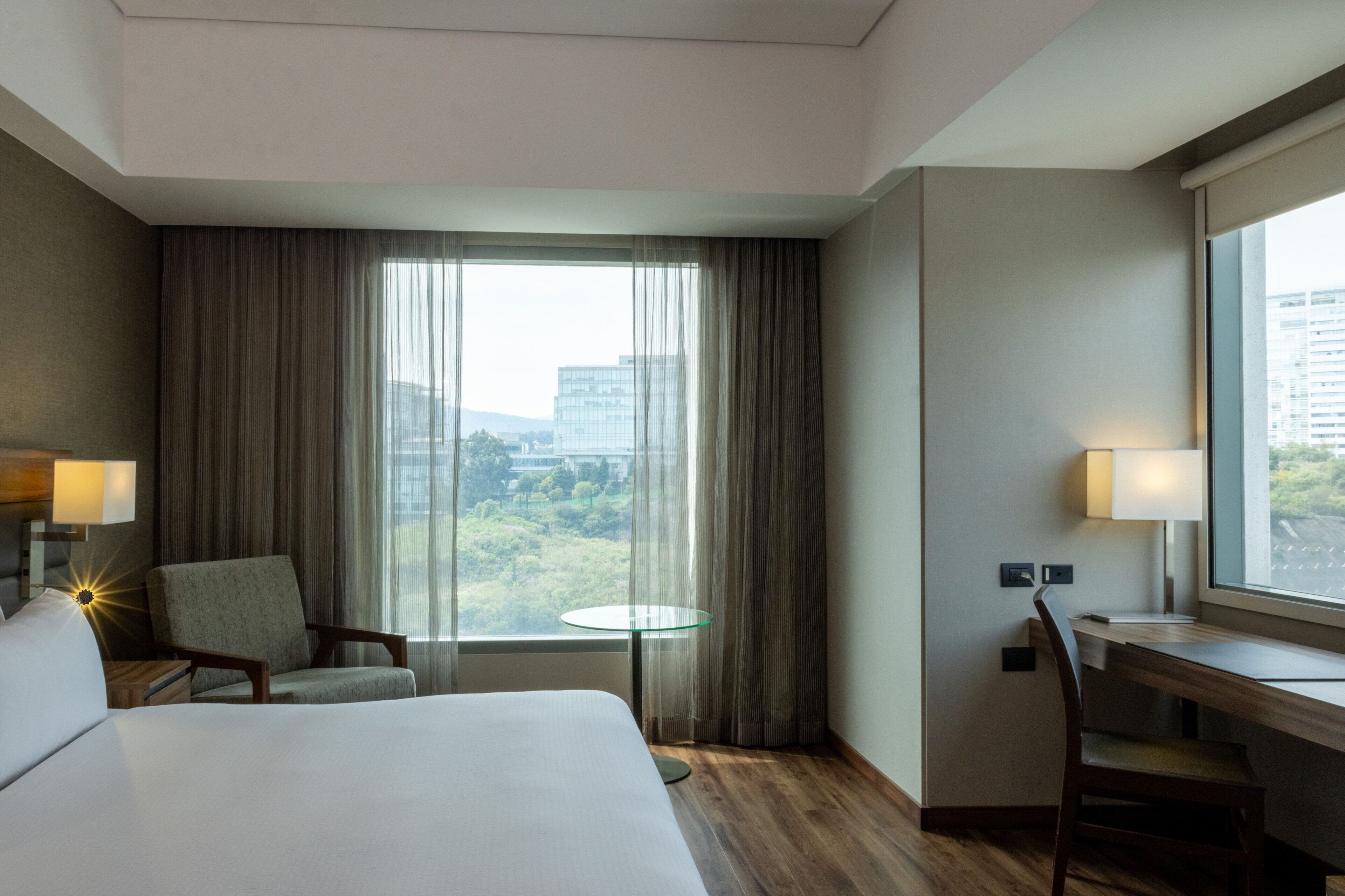A hotel room with a bed, a desk, a chair, a small table, and a window with curtains.