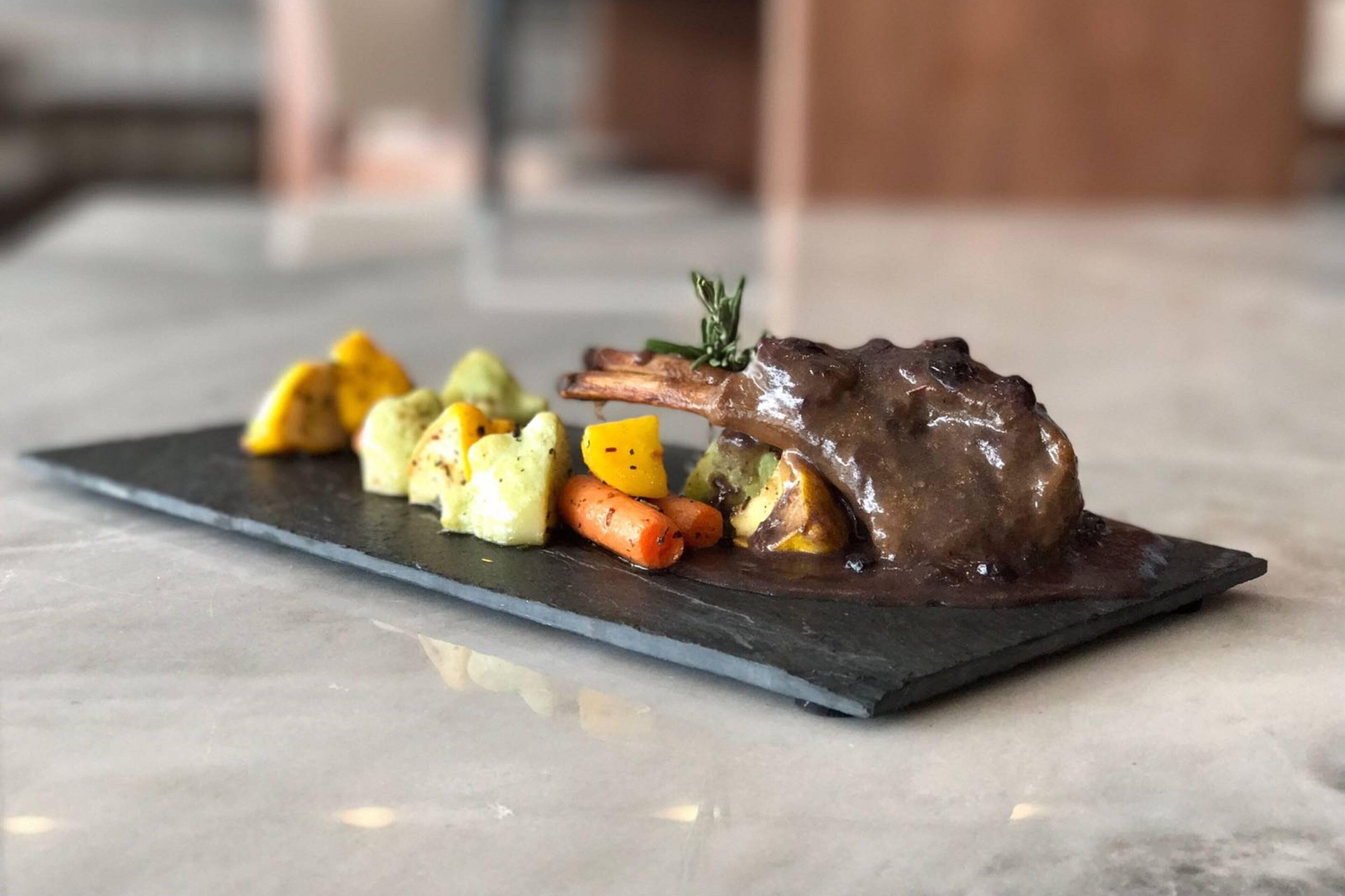 A plated dish with a lamb shank, roasted vegetables, and a dark sauce on a slate plate.