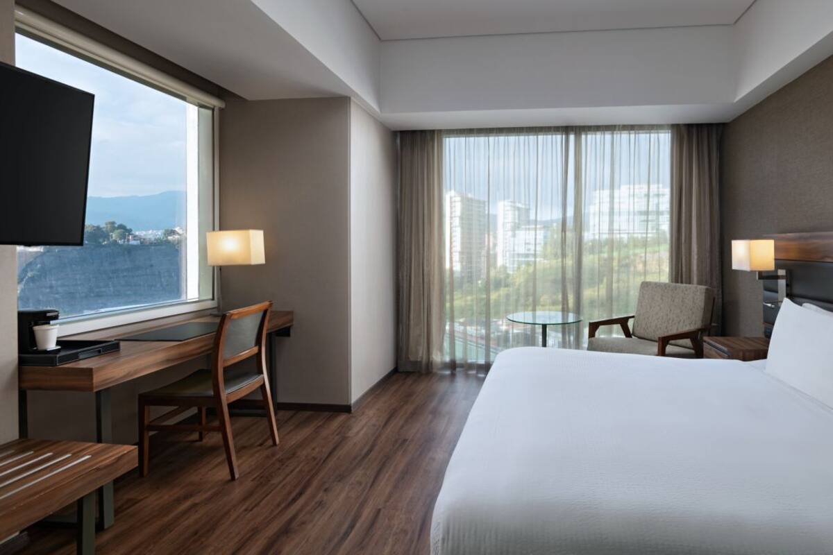 A hotel room with a large bed, a desk with a chair, a TV, and a view of the city through large windows.