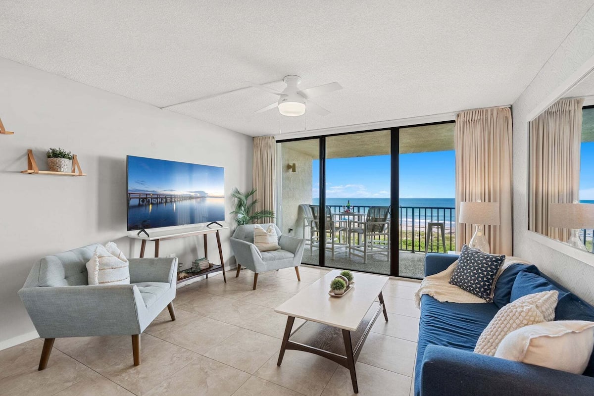 Een moderne woonkamer met een bank, fauteuil, salontafel, een televisie en een balkon met uitzicht op de oceaan.