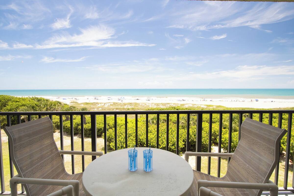 Een balkon met een tafel en stoelen, uitkijkend op een strand met mensen en parasols.