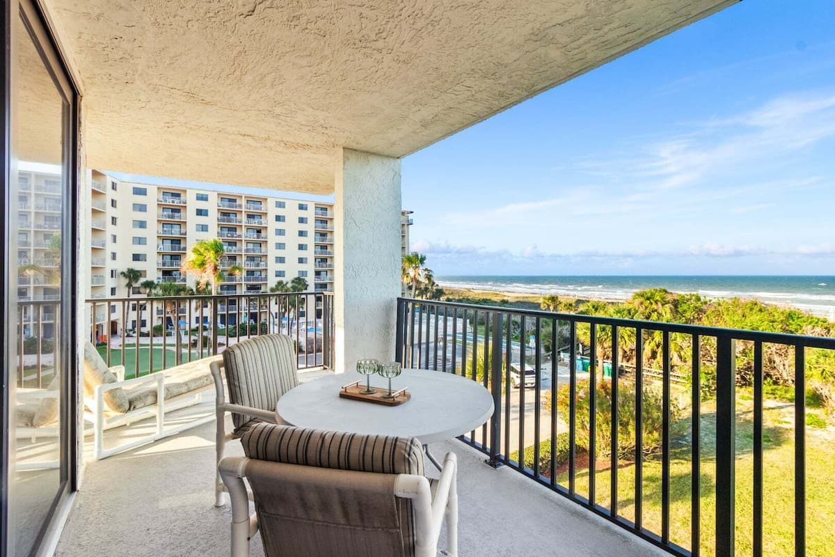 Een balkon met een tafel en stoelen, met uitzicht op een strand en een hoog gebouw.
