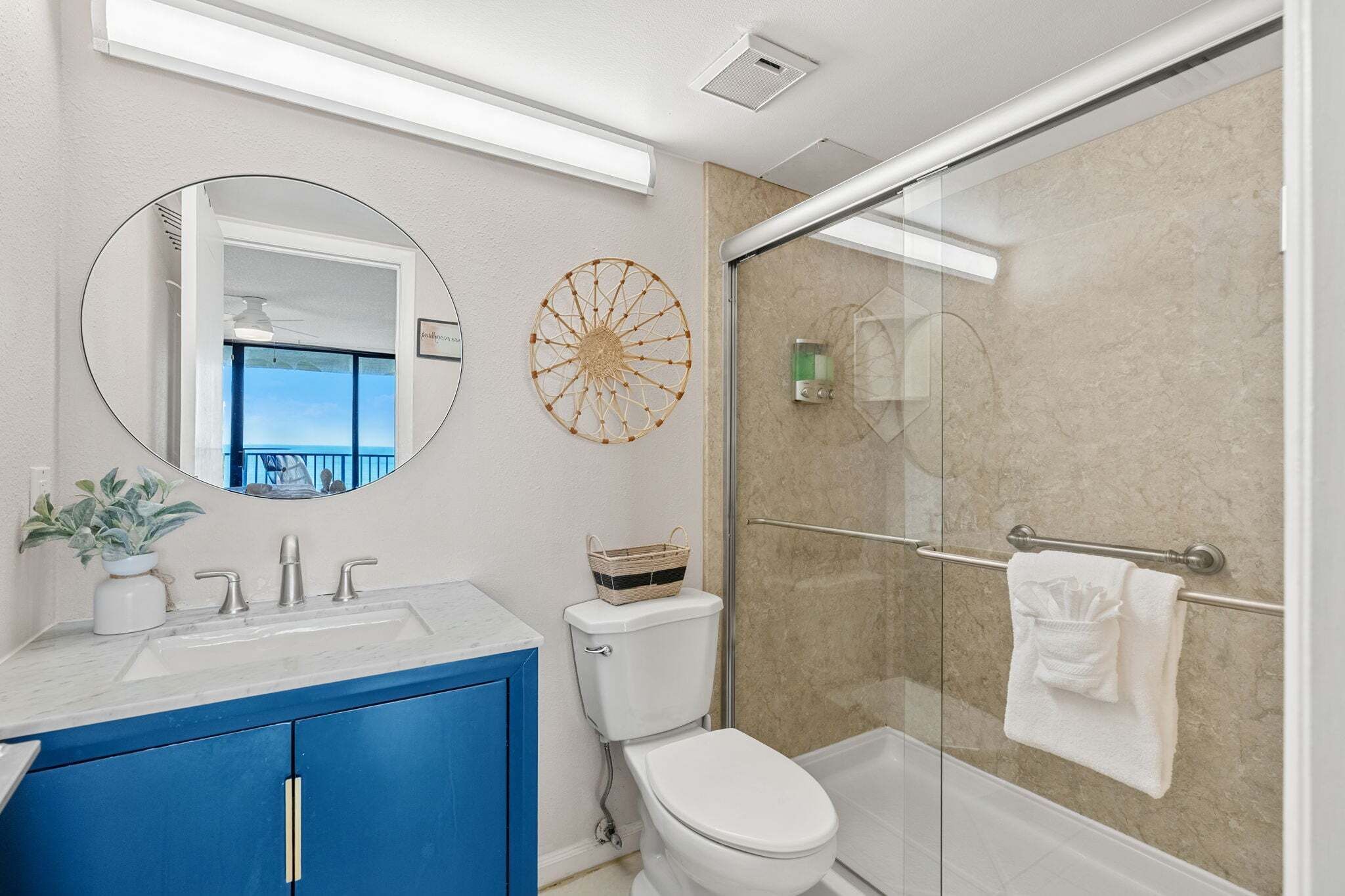 Un baño con cabina de ducha de vidrio, inodoro blanco, lavabo con mueble azul y espejo redondo.