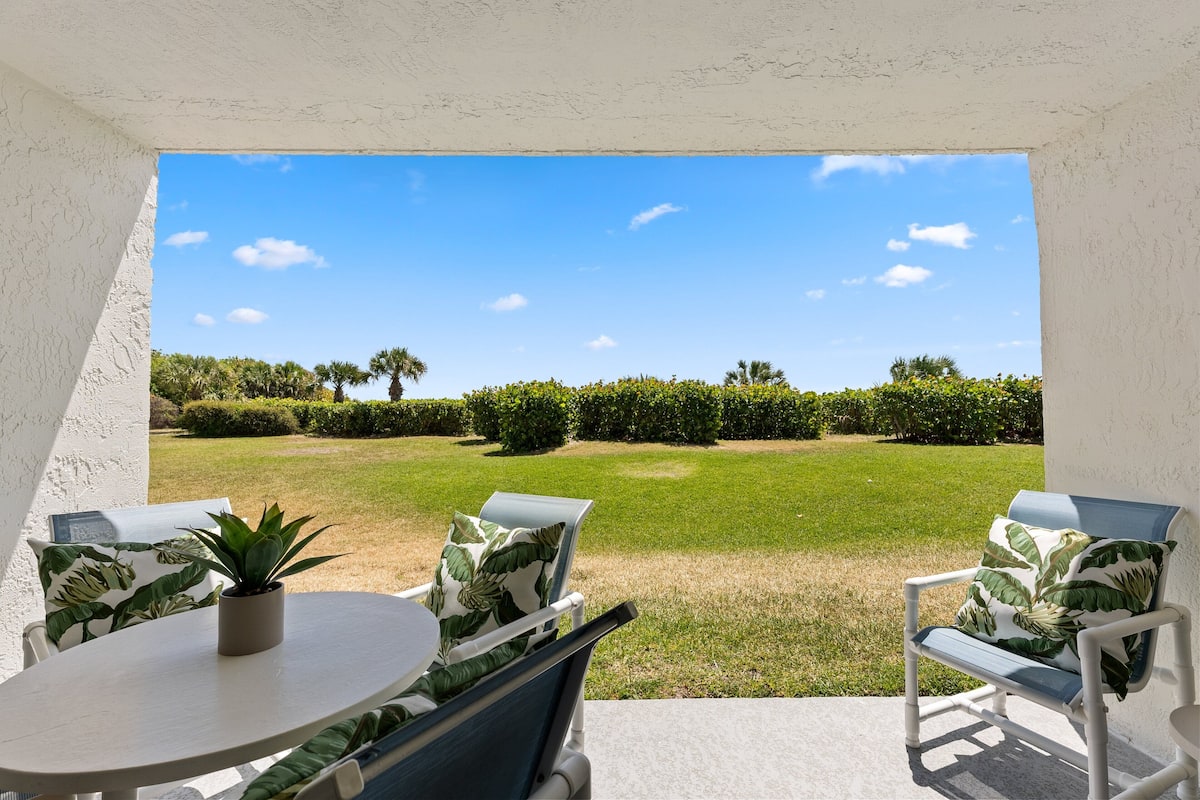 Een terras met een ronde tafel en twee stoelen, waarvan er een een plant heeft, met uitzicht op een verzorgd gazon en een helderblauwe lucht.