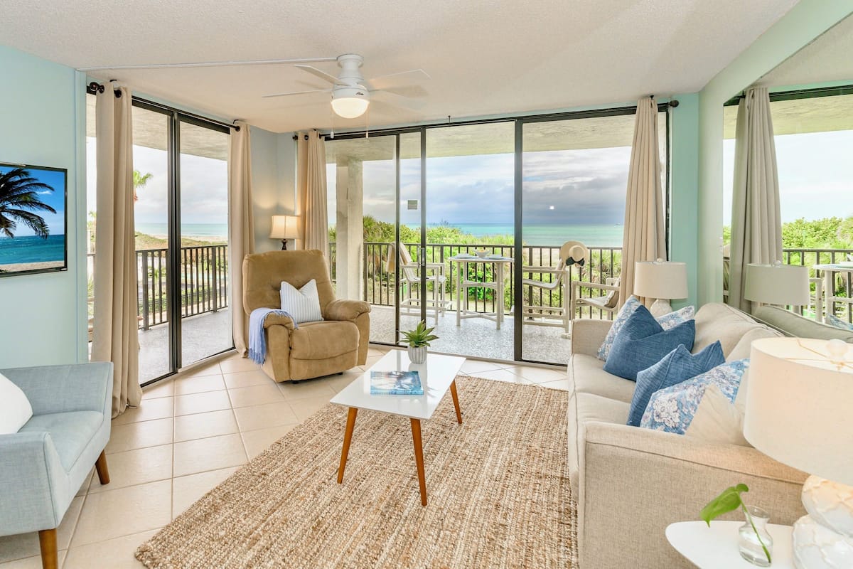 Een ruime woonkamer met een balkon, een plafondventilator en uitzicht op het strand.