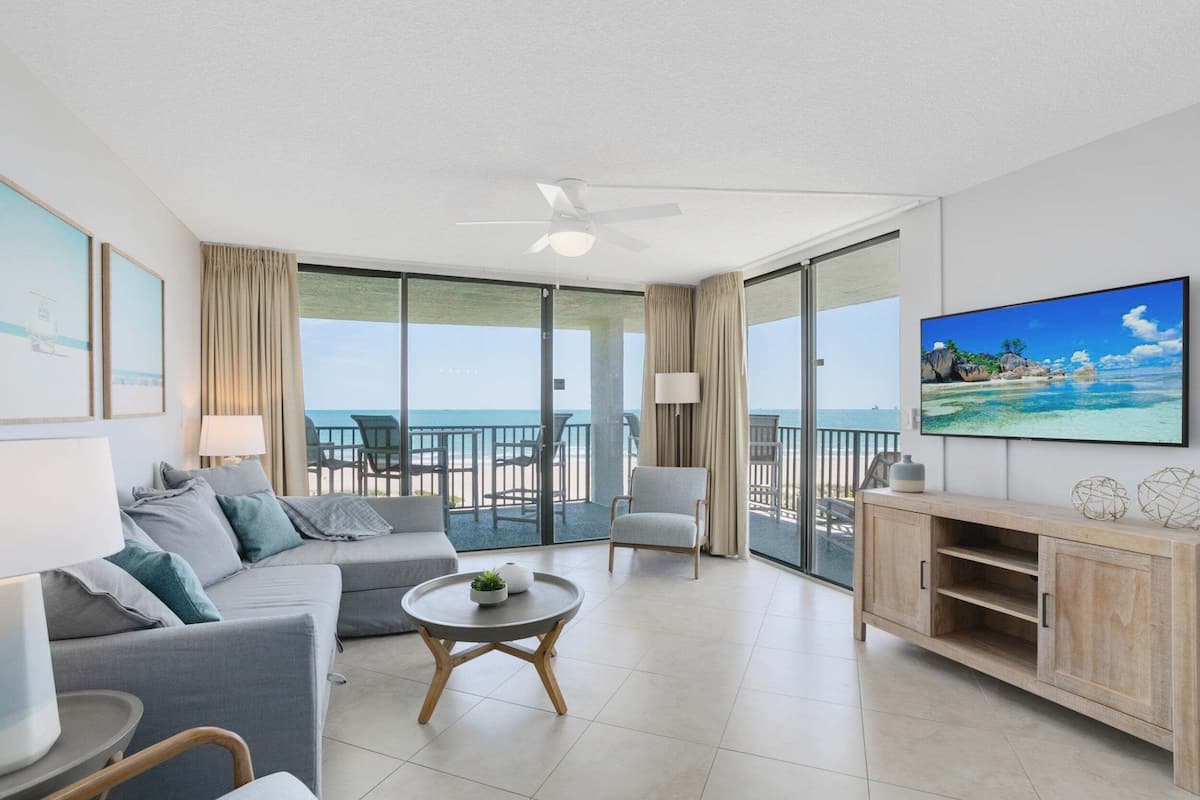 Een moderne woonkamer met een bank, fauteuil, salontafel en een televisie op een houten kast. Een balkon met uitzicht op de oceaan is zichtbaar door grote ramen.