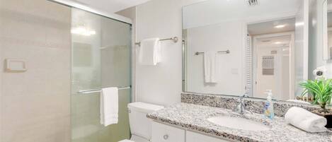A bathroom with a glass shower enclosure, a white toilet, a granite countertop sink, a large mirror, and a potted plant.