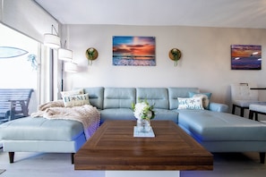 A modern living room with a light blue sofa, a wooden coffee table, and two wall-mounted paintings. - Cape Winds - Oceanfront Pool, Tennis & Sauna Condos near Port Canaveral (Cape Canaveral)