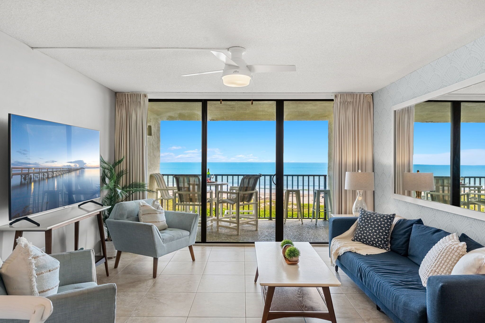 A living room with a TV, sofa, and coffee table, a balcony with chairs and a view of the ocean.